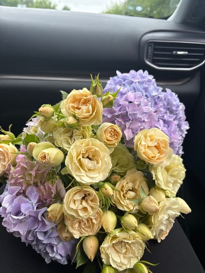A bouquet of yellow roses and purple hydrangeas is placed on the interior of a vehicle. The bouquet is in the foreground, with the roses predominantly yellow and peach-toned, and the hydrangeas a soft lavender. The background shows the dark interior of a car, including part of a dashboard and an air vent. The image appears to have been taken inside the vehicle.