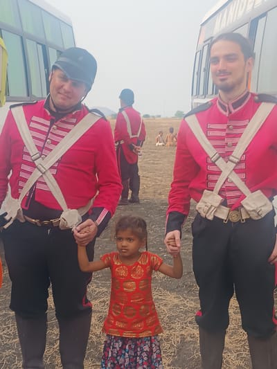 The image displays an outdoor scene in Kottalapalli, India, featuring three individuals in the immediate foreground. A young child, appearing female and of Indian descent, stands centrally, holding hands with two adult males positioned to her left and right. The child wears a red patterned top and a floral skirt. The adult male on the left is Caucasian, wears a black helmet, a red military-style jacket with white cross-straps and horizontal white bands, black trousers, and dark boots. The adult male on the right, also Caucasian, wears similar attire but without a helmet, his dark hair pulled back. Both adult males have small beige pouches attached to a belt with a gold buckle. In the midground, a third individual in a similar red uniform and black helmet is visible from behind, facing away from the viewer. Further in the background, several other individuals, some shirtless, are seated on the dry, dusty ground. Two large white buses are parked behind the foreground figures; one exhibits partial text "MANIB..." on its side. The ground surface is characterized by dry soil and sparse, light-colored stubble. The sky is bright and appears overcast or hazy.