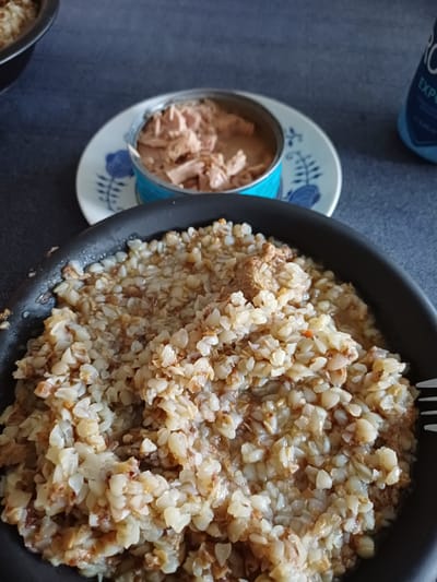 A close-up, eye-level shot shows a meal on a dark blue, textured surface, possibly a table or couch. In the foreground, a black, shallow pan is filled with cooked buckwheat groats mixed with pieces of meat. The groats are light brown and white, with some darker, browned bits. A portion of a white fork with metal tines is visible on the right edge of the pan. In the mid-ground, a small, white plate with a blue floral pattern holds an open can of tuna. The tuna is flaked and light pink. To the right of the plate, a blue plastic bottle with the partial text "EXPORT" is visible. The background is out of focus, showing more of the dark blue surface and a sliver of another dark bowl on the left. There are no people or identifiable landmarks of Ballerup, Denmark in the image.