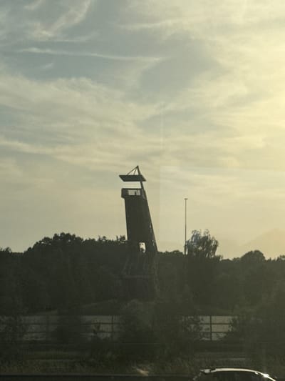 The image depicts the iconic sculpture "De Vonk" in Wuustwezel, Belgium, against a cloudy sky. The sculpture, which resembles a tower, is silhouetted against the bright, hazy sky. In the foreground, a line of trees and vegetation forms a dark band across the lower portion of the image. A fence is visible behind the trees. The overall scene suggests an outdoor environment, possibly a park or roadside, during the daytime. No people or vehicles are clearly visible.
