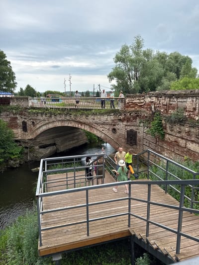 The image shows the bridge over the Turya River in Verkhoturye, Russia. The bridge is constructed of weathered red brick with an arched span. A wooden viewing platform with metal railings has been built next to the river. Several people are visible on the bridge and on the platform. On the bridge, a group of individuals are walking and standing, with trees and buildings visible in the background. On the platform, a family is gathered, including a man, a woman in a green dress, and a boy in a bright green shirt wearing a leg brace. The river flows beneath the bridge, with green vegetation growing along its banks. The sky is overcast.