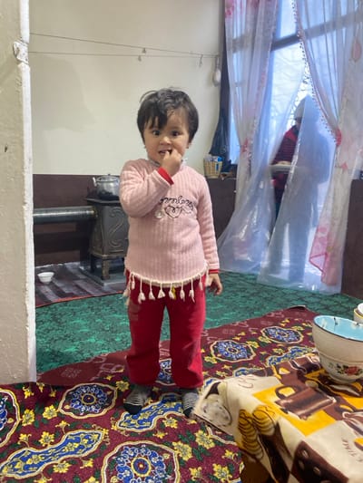 An interior scene in Akhtachi, Uzbekistan, features a young child standing center-left on a colorful patterned rug. The child, wearing a light pink knitted sweater with white tassels, red trousers, and striped socks, looks directly at the camera with wide eyes, fingers of the right hand positioned at the mouth. To the child's left, a dark metal stove with a horizontal pipe and an aluminum kettle on top rests on a green patterned rug. A small white bowl sits on the green rug nearby. The background features a light-colored wall with a darker lower section. To the child's right, a large window is covered by sheer white curtains with pink accents. Behind the curtains, a blurry figure, possibly an adult, wearing a red striped garment and a light-colored head covering, is partially visible, holding a flat object or tray. A dark piece of furniture with a wicker basket on top is located between the child and the window. In the immediate foreground on the right, a low table is covered with a yellow and brown patterned tablecloth displaying illustrations of coffee-related items, with several ceramic bowls, including a blue and white patterned one, placed on top.