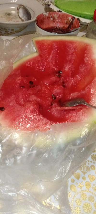 A close-up, overhead shot shows a slice of watermelon resting on a white plastic tablecloth. The watermelon is bright red with scattered black seeds and has a thin green rind. A silver fork is visible, partially submerged in the watermelon, suggesting it is being eaten. In the background, a small white bowl with a red floral pattern and a partially visible green plate can be seen. The overall scene appears to be an informal meal setting. The location is identified as Belgorodskoye, Kazakhstan.