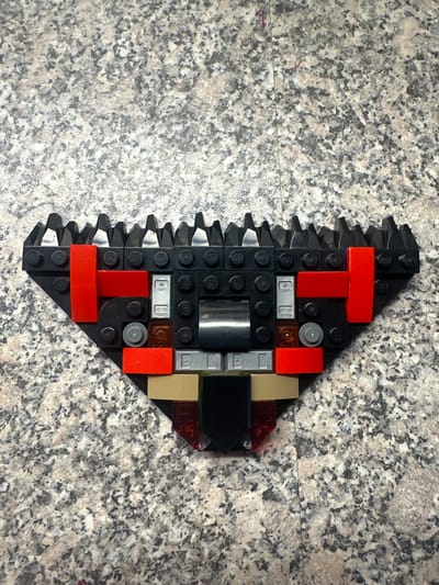 The image shows a black Lego build, resembling a stylized mouth or a gear, constructed on a speckled granite surface. The build features various black, red, silver, tan, and clear Lego pieces. The background is a textured, grey and white granite countertop. The image is an overhead shot. The city of Hüsen, Switzerland is not depicted in this image.