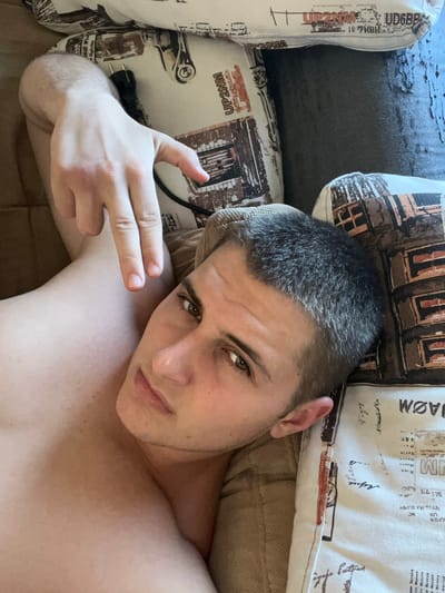 A young man with short, dark hair lies on his side, looking at the camera. He is shirtless, with his right arm bent and his hand held up in a gesture resembling a gun. He is lying on a sofa with patterned pillows. The pillows have text and images of buildings on them. A black electrical plug is visible on one of the pillows near his arm. The lighting suggests it is daytime, and the indoor setting appears relaxed. The man has a neutral expression. Text fragments visible on the pillows include "JAØM", "UP2NM", "SUPER", and "Hoara". The location is identified as Galiche, Bulgaria.
