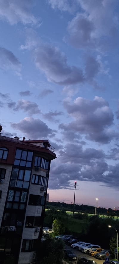 The image captures a twilight scene in Romashkovo, Russia, focusing on a modern residential building and the surrounding urban landscape. The sky is a deep blue with scattered grey clouds, suggesting the transition from day to night. In the foreground, the side of a multi-story building with a red tiled roof is visible. The building features numerous large windows, some with reflections of the sky, and air conditioning units installed on its facade.

Below the building, a parking lot is filled with various cars, including a yellow SUV and several dark-colored sedans and SUVs. The vehicles are parked in rows along a road. Beyond the parking lot, a slope covered with green foliage rises, dotted with trees. In the middle distance, a tall pole with a red light at its top stands out against the sky, likely a communication tower or part of a light signal. Further in the background, city lights and buildings are faintly visible, indicating the presence of a more developed urban area. The overall atmosphere is one of a residential district at dusk, with the subtle illumination of streetlights starting to appear.
