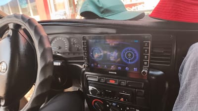 The image shows the interior of a car, likely a sedan, in the city of Mirzo Tursunzoda, Tajikistan. The dashboard features a modern infotainment system displaying the time as 08:05 and the date as 2026/06/29, with Russian text indicating it is a Monday. The screen shows music playback controls and a simulated speedometer indicating 0 km/h. To the left of the infotainment system, a leather-covered steering wheel with embossed patterns is visible, and beyond that, the car's instrument cluster with a speedometer and tachometer. On top of the dashboard, a green baseball cap and a red visor are placed. The overall condition of the car's interior suggests it is in use. The presence of a modern infotainment system in an older car model indicates a possible upgrade or aftermarket installation. The lighting suggests it is daytime, possibly morning, given the time displayed on the screen.