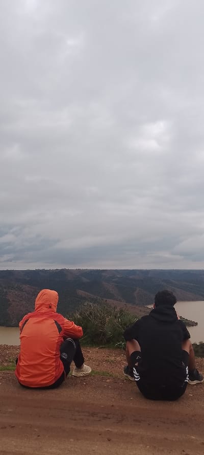 This image captures two individuals seated on a reddish-brown dirt surface, observed from behind, overlooking a panoramic landscape under an overcast sky, located near Aïn El Aouda, Morocco. The individual on the left wears an orange hooded jacket over black pants with white side stripes and light-toned sneakers. The individual on the right wears a black hooded sweatshirt with the hood down, revealing dark hair, black shorts with white side stripes, and dark athletic shoes. Both are facing away from the camera, towards a view that includes a wide, turbid brownish-tan body of water, presumably a river or reservoir, flowing through undulating hills. These hills are covered in sparse, dry, greenish-brown vegetation. The sky above is entirely obscured by dense, uniform grey clouds. The immediate foreground is bare earth with minor irregularities, and some low-lying green shrubs are visible between the individuals and the water.