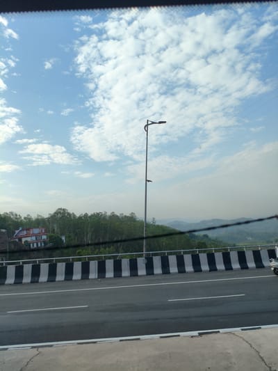 The image shows a highway in Bilaspur, India, on a cloudy day. The highway is divided into multiple lanes, and there is a concrete barrier with black and white stripes separating the lanes from the shoulder. A street light stands tall on the left side of the road. In the background, there are green hills covered with trees, and a few buildings are visible on the slopes. The sky is blue with scattered white clouds. The image is taken from a moving vehicle, as indicated by the blurred foreground and the slight motion blur in the image.