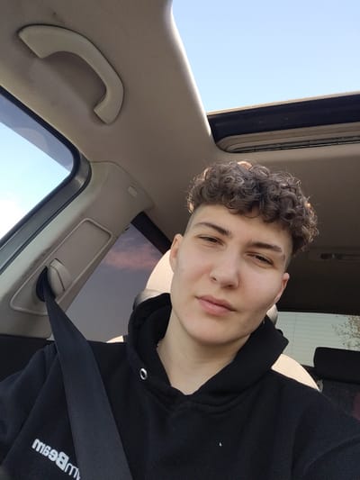 A young individual with dark, curly hair and shaved sides is captured in a front-facing selfie, seated in the driver's side of a car. They wear a black hooded sweatshirt with a white, reversed text on the front, which appears to read "mBeam" when unreversed, and are secured by a black seatbelt. The person looks calmly and directly at the camera with a neutral expression.
The car's interior features a light-colored headliner and pillar trim. A beige grab handle is visible above the driver's side window. An open sunroof reveals a bright blue sky, and through the car's windows, a clear blue sky is predominantly seen. A faint pinkish cloud is visible in the distance through the rear side window, indicating it is daytime. The setting is within a vehicle in Borisovo, Bulgaria.