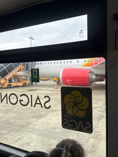 The image depicts an airport tarmac scene at Da Nang International Airport, located in Hòa Cường Ward, Da Nang, Vietnam, viewed through the window of a ground vehicle. A white passenger aircraft, featuring a large red engine cowling and red, orange, and yellow markings on its tail section, is parked on a concrete surface. Adjacent to the aircraft's fuselage is a yellow mobile airstair, which displays the text "SAIGON GROUND SERVICE." A green "Emergency Exit" sign with a running figure is affixed to the aircraft near the stairs. Additional yellow ground support vehicles and several orange traffic cones are visible across the tarmac. In the background, a tall light pole with multiple fixtures extends into an overcast, grey sky. Reflections on the window include the reversed word "SAIGON" and a rectangular sticker containing a yellow five-petal flower logo above the reversed text "SACS." The top of a person's head, exhibiting grey hair secured by a light-colored hair accessory, is visible in the bottom-center foreground, inside the viewing vehicle.