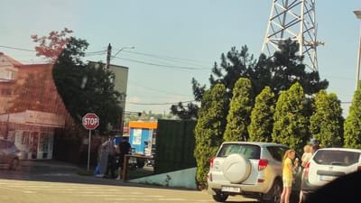 The image depicts a street scene in Ialoveni, Moldova, on a clear, sunny day. In the foreground, a silver SUV is parked on the right side of the frame, with its rear facing the viewer. Its license plate reads "PFS 254". To the right of this SUV, a white SUV is partially visible, with a license plate that appears to read "TBR". A young girl in a yellow top and shorts is standing next to the silver SUV, holding an ice cream cone. Behind the vehicles, several tall, conical green trees are arranged in a line.

In the middle ground, a street with white lane markings is visible. A red stop sign stands on a pole on the left side of the road. Two men are standing near the stop sign and a blue and orange structure. One man, wearing a patterned shirt and dark pants, is looking towards the right. The other man, in a dark shirt and shorts, is facing away from the camera. Adjacent to them is a green fence. Further back, a building with a red tiled roof is visible on the left, with a sign that reads "Prospetime". To the right of this building is a taller, grey building. A large metal tower structure is visible in the background, along with more trees and buildings in the distance, suggesting a suburban or town setting. Power lines crisscross the sky above the scene.