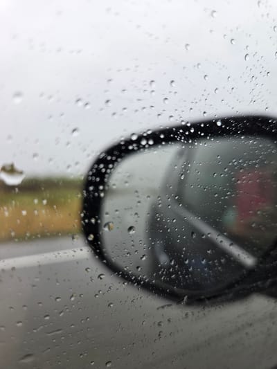 The image captures a close-up, out-of-focus view of numerous raindrops clinging to a glass surface, likely a car window. The droplets vary in size and shape, creating a textured foreground that blurs the elements behind them.
In the mid-ground, slightly out of focus, is the dark, oval-shaped frame of what appears to be a vehicle's side mirror or rearview mirror. The surface of the mirror itself reflects soft, indistinct colors, including hints of pink, green, and a muted yellow or brown, suggesting some distant objects or landscape are being mirrored. The light background around the mirror suggests an overcast sky or diffused daylight.
The scene depicts a moment during or immediately after rainfall, viewed from inside a vehicle. There is no specific activity taking place, but the image conveys the atmosphere of a rainy day from an interior perspective. No people are visible.
Notable details include the crispness of the individual water droplets contrasted with the soft, blurry background, emphasizing the glass as the primary focal point. The overall lighting is subtle and diffused. The provided location context is Бабстово, Russia, though no specific visual cues within the image confirm this location. There is no visible text.