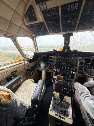 This image shows the interior cockpit of an old, possibly abandoned aircraft. The cockpit is weathered and shows signs of disuse. Moisture is visible on the windows. The control panels, filled with gauges, switches, and levers, show a worn and outdated design. The seats are padded with worn fabric. One person is partially visible in the right seat, wearing a grey top and beige pants. Another person has legs and feet visible on the bottom right and appears to be holding some kind of food. The South African registration of the aircraft, ZS-LYL, is noted on one of the panels. There is visible corrosion around the window frames, and portions of the window are obscured. Based on the aircraft registration and the context clues in the image, the location is likely in Kraaifontein, South Africa.