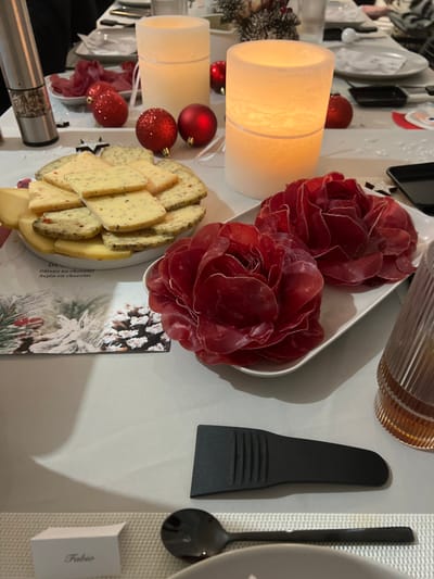 This is an overhead shot of a festive table setting, likely for a dinner or party. The scene is dominated by food and decorative elements, suggesting a celebratory occasion, possibly during the winter holidays.
In the center of the image, two platters hold appetizers. One platter contains thinly sliced red cured meat, possibly bresaola or prosciutto, artfully arranged into two large rose-like mounds. Adjacent to it is a platter with various types of cheese, cut into triangular and rectangular pieces, some appearing to be infused with herbs or spices.
The table is decorated with two tall, cylindrical white candles, casting a warm glow, and several glossy and glittery red spherical Christmas ornaments. A small sprig of faux evergreen with white berries is also visible. Beneath the meat platter, a card with a winter motif featuring pinecones, evergreens, and red berries is partially visible. This card also has French text, including "Gateau au chocolat" (chocolate cake) and "Sapin en chocolat" (chocolate Christmas tree), suggesting a dessert menu or theme.
Other objects on the table include a black serving spatula, a black spoon, a ribbed transparent glass, a silver and clear pepper grinder, and a white placemat. A folded white card with the name "Fabio" printed on it serves as a place card. In the background, partially visible plates and what appears to be a Santa figurine further contribute to the festive atmosphere. The setting appears to be indoors, consistent with a home in Bellevue, Switzerland, during a special occasion.