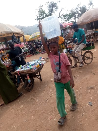 This image depicts an active outdoor market scene in Kpak, Nigeria, characterized by a light brown dirt ground under an overcast sky. In the foreground, a young individual wearing a pink t-shirt with a blue graphic, green track pants, and grey slide sandals faces forward, looking directly at the viewer. This individual balances a large, clear plastic bucket on their head, supported by a multi-colored woven head-pad, with their right arm raised to steady the container. A small, patterned cross-body bag is worn over their left shoulder.
To the left of the central figure, a man in a black long-sleeved shirt stands behind a wooden wheelbarrow functioning as a vendor's stall, displaying a variety of packaged goods, predominantly in white, blue, and green packaging. To the right, a man wearing a teal t-shirt, light cargo shorts, and a dark knit cap is seated on a black bicycle, which is modified with a front basket and a rear rack holding a green plastic crate containing several dark-colored bottles. He faces forward, appearing to be in motion or paused.
The background reveals a densely populated market area with numerous stalls shaded by assorted tarpaulins and umbrellas (tan, yellow, red), indistinct figures, and distant green hills under a bright, diffuse sky.