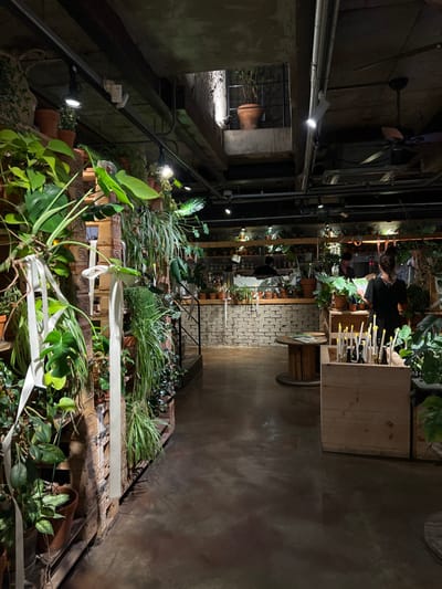 The scene depicts the interior of a plant-focused commercial establishment, likely a plant cafe or shop, located in 마포구, South Korea. The environment is dimly lit with an exposed concrete ceiling featuring visible pipes, electrical conduits, track lighting, and ceiling-mounted fans.
Along the left side, a prominent vertical display of various potted plants, including large-leafed and trailing species, is arranged on stacked wooden pallets. Several plants have white ribbons tied to them. The floor is dark, polished concrete.
In the background, a service counter with a light-colored brick facade is visible. Two individuals in dark attire are present behind this counter; one is centrally positioned, facing the counter, while the other is partially obscured, performing an action. Shelves above the counter hold numerous small potted plants.
In the mid-ground to the right, a person with dark hair, wearing a dark top, stands facing a light-colored wooden display box filled with various plant tools or accessories. This individual is observed examining an item in their hands. A round wooden cable spool serves as a small table in the mid-ground. A black-railed staircase ascends on the left, leading to an upper mezzanine level where a large terracotta pot containing a tall, slender plant is visible.