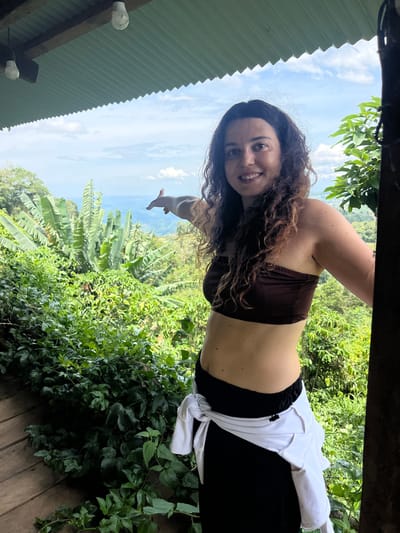 A woman with curly brown hair stands on a wooden deck, pointing to the right with her left hand. She wears a brown tube top, black pants, and a white jacket tied around her waist. The background shows lush green vegetation, including banana trees, and a hazy landscape with distant hills under a cloudy sky. Two light bulbs hang from the corrugated metal ceiling of an overhang. The location is described as the city of Son Dien, Vietnam, though this specific location within Son Dien is not discernable from the image.