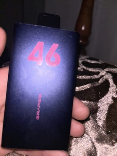 The image shows a close-up of a rectangular, dark blue object, possibly a vape device or a small power bank, held in a person's hand. The object has the number "46" printed in bright pink on its surface, and below that, the word "GRAPES" is also printed in pink. The background is dark and appears to be an indoor setting, with a textured, possibly upholstered surface visible. There is no indication of the location being in Paraul, Russia, or any other specific city.