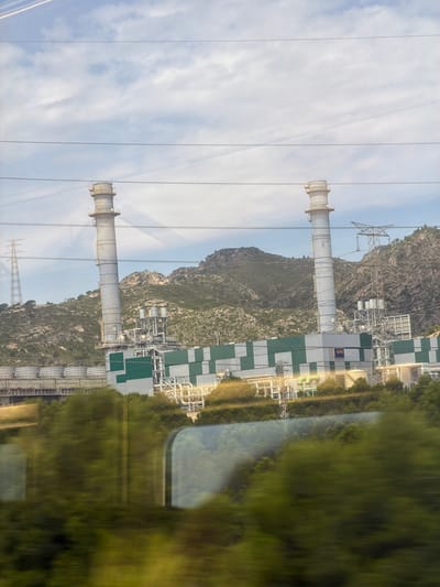 This is a medium-distance, eye-level, slightly angled, landscape orientation photograph taken from inside a moving vehicle, likely a train, as suggested by the blurred green foliage and reflections in the foreground that appear in the windows. The image depicts a large industrial facility situated in front of a rugged, sparsely vegetated mountain range under a partly cloudy sky.
Dominating the center of the frame are two tall, slender industrial smokestacks. The one on the left is light gray, and the one on the right is also light gray, but appears slightly shorter and more weathered. Both smokestacks have some visible piping and structural elements near their tops.
Below the smokestacks, a long, rectangular industrial building stretches horizontally. This building has a distinctive pattern of alternating light gray and dark green rectangular panels. A blue sign with the orange letters "ALPLA" is visible on the front of this building. The building is surrounded by a complex network of interconnected pipes, tanks, and smaller structures, indicative of a processing plant. To the left of the main building, a series of large cylindrical tanks with white tops are visible, also part of the industrial complex.
In the background, the mountain range rises steeply. The slopes are covered in scattered green vegetation and rocky outcrops. The sky above is a pale blue with scattered, wispy white clouds. Several power lines and a tall transmission tower can be seen in the distance, further indicating an industrialized area.
The foreground consists of blurred green trees and bushes, their motion captured by the camera due to the vehicle's movement. Reflections of the interior of the vehicle, particularly the curved shapes of windows, are visible within this blurred foliage.
The overall scene suggests an industrial area, possibly a power plant or chemical facility, located in a somewhat remote or semi-rural setting near a mountainous region. Given the "ALPLA" signage, this facility could be related to plastic packaging production. This specific industrial complex is identifiable as the ALPLA factory located in the municipality of El Morell, within the comarca of Tarragonès, in the province of Tarragona, Catalonia, Spain.