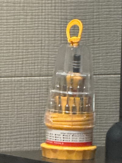 A plastic container holds a set of interchangeable screwdriver bits. The container is cylindrical and transparent, with a yellow base and a yellow loop at the top for hanging. The bits are visible inside the container, appearing to be made of metal. The container is positioned on a dark surface, with a textured gray wall in the background. There are no people or animals in the image, and no discernable actions are taking place. The image is focused on the screwdriver bit set.