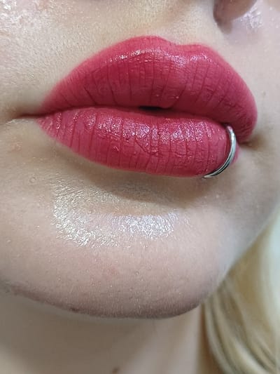 This image presents a close-up, partial view of an individual's face, specifically focusing on the mouth and lower facial area. The scene is identified as being located in 서대문구, South Korea. The subject's lips are coated with a deep red, matte lipstick, displaying visible fine lines and texture. A silver-colored, C-shaped or open hoop piercing is present on the lower lip, positioned towards the viewer's right side. The surrounding skin below the lips is fair-toned, exhibiting visible pores and a subtle, natural sheen. A small section of light-colored hair is visible at the far right edge of the frame. No specific actions or interactions are depicted beyond a static pose, and the immediate background environment is not discernable due to the close crop.