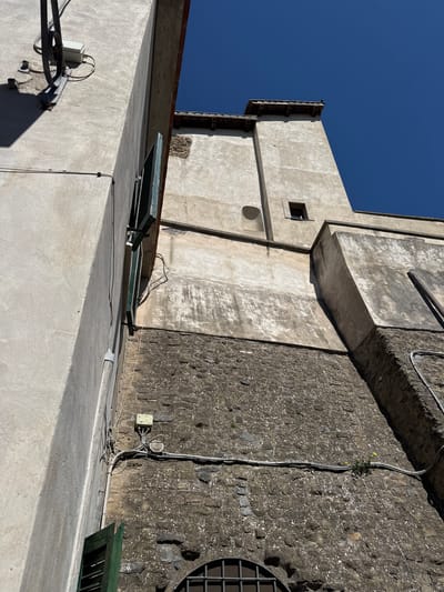 A low-angle shot captures building exteriors under a clear blue sky in Castelnuovo di Porto, Italy. The buildings exhibit a mix of materials, including concrete and stone walls. Electrical conduits are visible along the walls. The shot appears to be taken from a rooftop or elevated position.