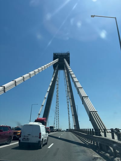 The media file shows a view from a vehicle traveling on a suspension bridge, likely during the day given the bright sunlight. The sky is a clear, vibrant blue, suggesting good weather. The perspective is from the passenger side of the vehicle, looking upwards and forward.

The prominent subject is the massive structure of the suspension bridge itself. Thick, parallel cables, encased in what appear to be white protective sheathing, ascend from the bridge deck towards the sky. These cables are supported by a tall, dark concrete pylon that dominates the center of the frame. The roadway of the bridge is visible, with lane markings and a concrete barrier separating the traffic lanes from the edge.

Several vehicles are on the bridge, indicating traffic. In the foreground, a white van is clearly visible, with its rear facing the viewer. To its left, a red SUV and a dark-colored car are also present. Further down the bridge, more vehicles, including trucks and vans, are seen in a line, suggesting a potential traffic jam or slow-moving traffic.

The overall scene conveys a sense of scale and engineering marvel, with the imposing bridge structure set against the vast expanse of the sky. The presence of vehicles indicates this is a functional transportation route.

Visible text on the white van's license plate reads "CT 73 OMV". There are also some circular stickers with numbers and symbols on the rear of the van. A small bird can be seen flying in the sky.

The location is Lazu, Romania, which is consistent with the presence of a large bridge. The time of day appears to be mid-morning or early afternoon, due to the strong sunlight and clear skies. There are no explicit emotional cues conveyed, other than the potentially frustrating experience of being in traffic.
