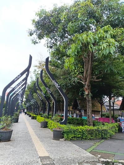 The image displays a pedestrian pathway in thành phố Biên Hòa, Vietnam, under an overcast sky. The pathway, paved with grey aggregate material and featuring a central yellow tactile guidance strip, extends from the foreground toward the background. Along the left side of the pathway, a linear series of approximately fifteen black, upward-curving arch-like structures stands, each inscribed with white Arabic calligraphy including phrases such as "الرحمن" (Ar-Rahman), "القدوس" (Al-Quddus), "السلام" (As-Salam), and "الرحيم" (Ar-Rahim). Dark grey potted plants are regularly spaced between these structures and a parallel low, manicured green hedge on the right. Beyond the hedge, a dense canopy of large trees with green foliage partially obscures several small background structures featuring rustic, possibly thatched or tiled, roofs, suggesting vendor stalls or small eateries. Some colorful outdoor seating, including pink and turquoise items, is visible near these structures. Multiple individuals are observed walking on the distant section of the pathway, moving away from the viewer. An additional person is partially visible on the far right, adjacent to the background structures.