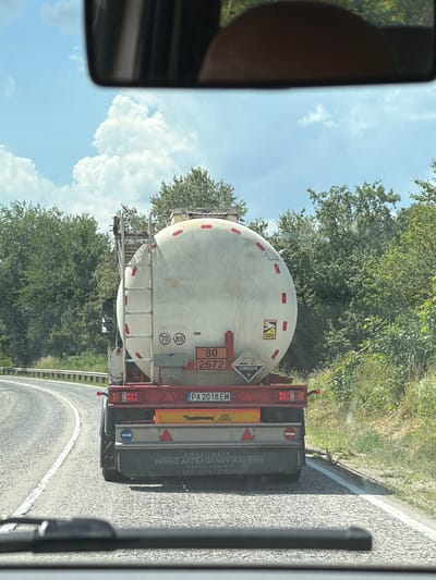 A tanker truck is driving on a paved road. The truck is white with red markings and is carrying a large, white, spherical tank. There are hazard labels on the side of the tank, including one that says "CORROSIVE" and another with a skull and crossbones. The truck also has license plates. The road is bordered by trees and greenery. The sky is blue with some clouds, suggesting it is daytime and the weather is fair. The image is taken from inside another vehicle, likely looking in the side mirror or out the passenger window. There is text on the side of the truck's trailer, some of which is in Greek. The text on the rear of the trailer appears to be company information, possibly including a name and phone number. The scene appears to be in a rural or semi-rural area.