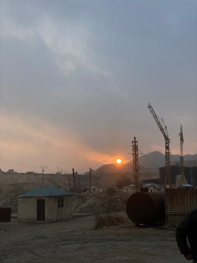 A dusty, hazy landscape features construction cranes silhouetted against an orange sunset. Industrial structures and small buildings dot a rugged terrain. A metal cylinder lies in the foreground near a concrete platform. The sky fades from light gray to an orange hue around the sun. Power lines are visible near the horizon.