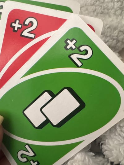 The image shows a close-up view of a hand holding three Uno cards. The visible cards are: a red "+2" card, a green "+2" card, and a green "draw two" card. The background is a blurry, light-colored, textured surface, possibly a blanket or other soft material. There is no evidence to indicate the location of Glojene, Bulgaria.