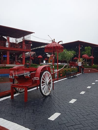 The scene depicts an outdoor leisure area in Tanjungarum, Indonesia, under an overcast sky, with visible moisture on surfaces indicating recent precipitation. The foreground features a stationary, decorative, two-wheeled red horse-drawn buggy with white spoked wheels, positioned on a wet, dark gray paved surface patterned with rectangular pavers laid in a herringbone style, marked by intermittent white dashed lines. To the left, a multi-story red building with dark roofs and open balconies houses what appears to be a dining or seating area. To the right, a single-story red open-air structure with a dark roof stands beside cultivated green foliage, including small trees and red flowering bushes. Numerous red Balinese-style ceremonial umbrellas (payung) are dispersed throughout the area, some freestanding, others integrated into the structures. String lights are visible under overhead coverings. A black ornamental metal bench is situated to the right of the central buggy. No individuals are present in the image.