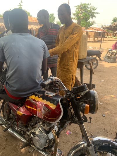 In Gobbiya, Nigeria, a group of men are gathered outdoors near a dirt road, with several buildings and trees visible in the background. The focus of the image is a motorcycle parked in the foreground. A man wearing a yellow traditional outfit is standing next to the motorcycle, appearing to interact with another man who is seated on the motorcycle, facing away from the camera and wearing a grey t-shirt and red shorts. Another man in a striped shirt is also present, looking towards the interaction. In the background, a rough wooden table holds a large plastic container. The scene depicts a casual gathering in what appears to be a rural or semi-urban setting.