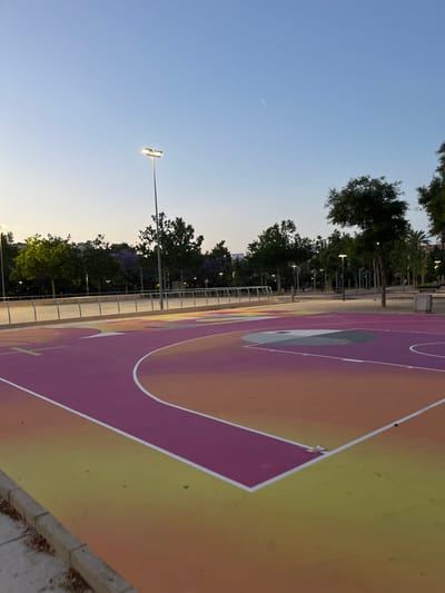 The media file displays an outdoor, modern, and brightly colored basketball court in Torrent, Spain. The court is predominantly painted in vibrant shades of purple, orange, and yellow, with crisp white lines delineating playing areas, including a key and a segment of the three-point arc. A circular gray and white design is visible within the purple section of the court.
No people are present, and no activity is currently taking place, though the court is clearly designed for sports. The scene is set in what appears to be late afternoon or early evening, indicated by the soft, clear blue sky overhead and the illuminated streetlights lining the perimeter. A faint contrail is visible in the upper left of the sky.
The court is bordered by a metal railing, beyond which a row of mature trees stands, some exhibiting green foliage and others with purple blossoms, possibly jacaranda. Several modern streetlights, each with multiple luminous heads, are visible among the trees, casting a warm glow. A paved path or sidewalk runs alongside the court. The general atmosphere is tranquil and ready for use. No visible text is present in the image.