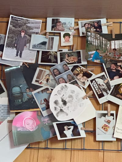 A vertical photo display board, constructed from a light brown wooden surface with an overlaying dark blue grid, presents a dense arrangement of personal photographs. Approximately 30-40 images of various sizes, predominantly small, instant-print style with white borders, are affixed to the grid using numerous small wooden clothespins. The scene is located in 3-Y Uchastok, Russia.
Notable photographs include a large vertical print in the upper-left depicting an older man in a dark coat and hat standing in a snowy landscape with a building; this photo bears the Russian text "Солотча" and the year "2013." Adjacent images feature a close-up portrait of a young woman and a beach scene. Further right, a group of people is shown gathered around a table with food, and another larger print displays individuals seated at a wooden table in a wooded outdoor setting under a tarp.
Central to the display is a prominent circular white paper bearing multiple dark gray fingerprints or smudges. Surrounding this are various photos portraying individuals and groups, including young women taking selfies, casual portraits, and scenes depicting social gatherings. Specific images include a vintage-style photo of a baby in a wicker pram, a smiling young woman in a pink hat, a woman posing on a beach, an architectural interior, and a close-up of miniature desserts. Partially visible Russian text, including "ФАТАЛИСТ" (Fatalist) and a handwritten note starting with "Таня" (Tanya), appear on papers attached to the lower portion of the board. The collection collectively documents personal memories and events.