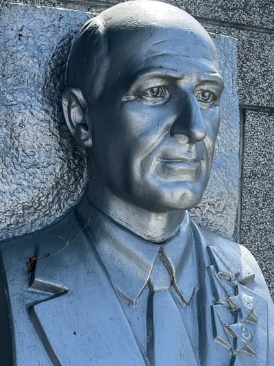 A silver-toned, bas-relief bust of a man is prominently displayed against a textured, grey background. The man appears to be middle-aged, with deep-set eyes, a prominent nose, and a stern expression. He is depicted wearing a suit with a tie and what appears to be a medal or decoration on his lapel. The bust is slightly weathered, with visible textures and some debris on its surface, including a small twig on the shoulder. The background is a rough, stone-like material, characteristic of outdoor monuments or architectural elements. This monument is located in the city of Takhtamukai, Russia.