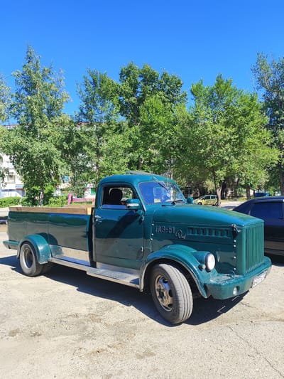 A dark green, classic pickup truck, identified by the "ГАЗ-51" emblem on its grille, is parked outdoors on a paved surface under a clear blue sky. The truck has a wooden-lined bed and prominent chrome trim along its side. Behind the truck, a line of leafy green trees forms a backdrop, with a building and other vehicles, including a yellow car, visible in the distance. The scene appears to be in an urban environment, likely Slavgorod, Russia, given the context. No people are visible in the image.