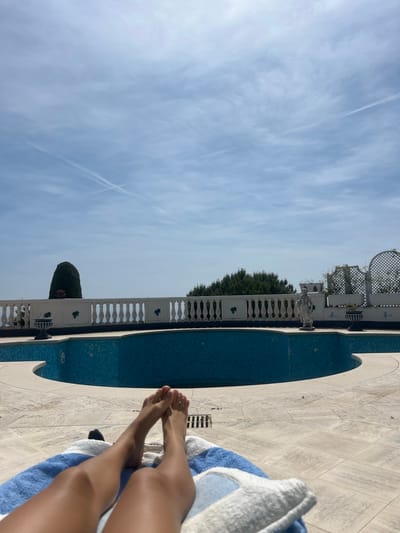 The image displays a serene, sun-drenched outdoor scene, likely in Èze, France. The perspective is from a person relaxing, with their bare legs and feet visible in the foreground, resting on a blue and white striped towel.

The person is positioned on a light-colored, square-tiled patio or pool deck. Immediately adjacent is a curved swimming pool, distinctive for its intricate blue mosaic tiling. Beyond the pool, a elegant white balustrade with classical designs extends across the mid-ground. This balustrade is adorned with decorative urns, a small statue, and sections featuring white trellises intertwined with green foliage.

Further in the background, a line of dark green trees and bushes adds to the lush landscape. The upper portion of the image is dominated by a expansive, light blue sky, dotted with soft, wispy white clouds and faint airplane contrails, suggesting clear weather. The overall ambiance is one of peace and leisure, typical of a luxurious vacation setting on a bright, sunny day. No visible text is present in the image.