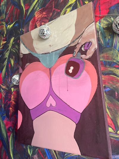 The image shows a close-up of a painting on a canvas. The painting depicts a stylized representation of a person's lower torso and upper thighs, rendered in bold, flat colors. The subjects are a pair of buttocks in shades of pink and orange, with a purple bra-like garment. Above this, a hand is shown with a purple nail polish bottle, with a drop of polish falling onto the buttocks. The background of the painting is a dark, textured color. Attached to the canvas are two small disco balls, one near the top and another on the left side. The painting is positioned in front of a vibrant, tropical-themed backdrop, which appears to be wallpaper or fabric, featuring large leaves in various shades of green, red, and yellow. The lighting suggests an indoor environment. There is no identifiable location in the city of Bucha, Ukraine, present in this image.