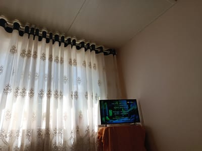 The image shows a television screen displaying credits, likely from a movie or show, positioned on a table covered with an orange cloth. To the left, sheer white curtains with a decorative pattern are drawn across a window, allowing sunlight to filter through. The room appears to be a domestic interior, possibly a living room, with plain walls and a ceiling. The location is identified as Shahrisabz City, Uzbekistan.
