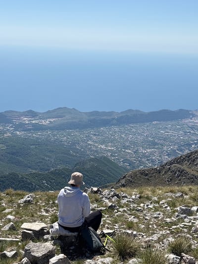 The image depicts a person sitting on a rocky, grassy cliff overlooking Mali Mikulići, Montenegro. The person wears a light gray hooded sweatshirt and a tan hat. A dark backpack rests behind them, with yellow trekking poles attached. The background shows a vast landscape, including mountains and a city below, under a clear blue sky. The perspective is from above, looking down at the person and the surrounding environment.
