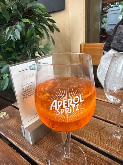 A glass of Aperol Spritz sits on a wooden table, next to a menu card, likely at an outdoor cafe or restaurant. The menu card lists "Aperol Spritz," "Sarti Spritz," "Hugo Spritz," "Limoncello Spritz," "Crodino Spritz," and "Pink Grapefruit," suggesting a range of spritz cocktails are offered. Another wine glass is visible in the upper part of the photo. A green plant is visible in the lower left corner, suggesting a natural element to the outdoor setting. The scene is located in Drammen, Norway.