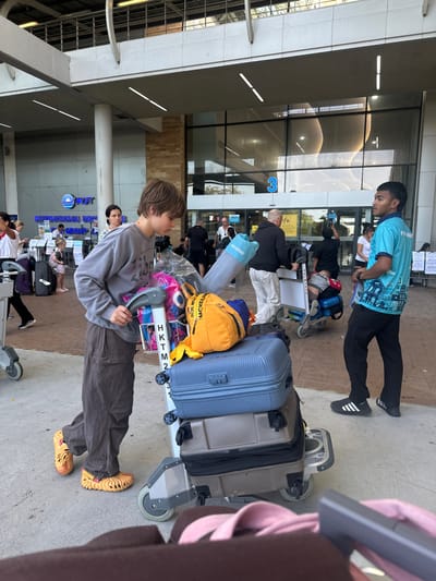 At an airport terminal, a young person in a gray sweatshirt, brown pants, and yellow shoes pushes a luggage cart loaded with suitcases, a yellow backpack, and other items. A man in a turquoise shirt and black pants stands beside a line of people with their luggage carts waiting to be processed. The airport exterior has large windows and signage.