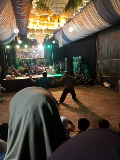 The image captures a vibrant cultural performance taking place at night or in the evening within a tent-like structure, likely in Kecamatan Cimaung, Indonesia. In the foreground, the back of a person's head, covered by a light-colored hijab, indicates a spectator's perspective. Several other audience members are partially visible in the lower frame, seated on the ground.

At the center, two men are actively engaged in a performance or demonstration on a dirt ground area. One man, wearing a blue shirt and dark pants, has his arms raised, while the other, in a brown shirt and dark pants, strikes a wide stance, casting a noticeable shadow. They appear to be performing a traditional dance or martial arts.

Behind the performers, an elevated stage hosts a group of musicians seated with various traditional instruments, including drums and what appear to be stringed instruments. Above them, a large banner displays text, partially readable as "LINGKUNG SENI PRABU..." and "NGABAYAN CIKAL...". The stage is lit by spotlights, including distinct green and red lights, against a black backdrop. To the right of the stage, stacks of green plastic chairs are visible, along with more audience members seated on the ground.

Overhead, the tent ceiling is draped with white, blue, and striped fabric, creating an indoor-like setting. Ornate, multi-layered chandeliers adorned with warm string lights hang from the ceiling, along with several white spherical lanterns, contributing to a festive atmosphere. The overall scene depicts a community event focused on traditional arts and cultural expression.