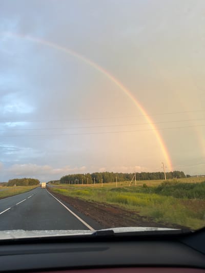 The image depicts a road with a double rainbow overhead, likely taken from inside a vehicle. The road stretches into the distance, bordered by green fields and a dense line of trees. Several utility poles are visible along the side of the road. A white truck is seen driving on the road ahead. The sky is overcast with the sun's light casting a warm glow, illuminating the rainbow. The scene is set in a rural environment, specifically in the city of Таймурзино, Russia, during daylight hours.