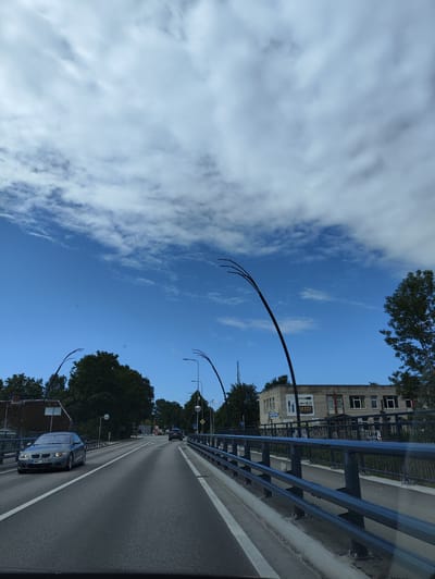 The image shows a street view in Salacgrīva, Latvia, captured from a moving vehicle. The scene features a two-lane road, with a grey BMW sedan in the foreground on the left lane, license plate MM-909. Another vehicle is visible further down the road in the right lane. Both lanes are bordered by a dark metal railing.
On the right side of the road, a two-story brick building with multiple windows is visible, with a sign that appears to read "HARDY". Trees and other buildings can be seen in the background. The sky is a bright blue with scattered white clouds. The image conveys a sense of travel through a suburban or semi-urban area on a clear day.