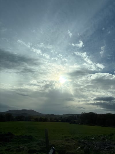 The image depicts a landscape in Shelve, United Kingdom. The scene is dominated by a cloudy sky, with the sun shining through a break in the clouds. Below the sky is a green field, edged by a tree line and a hill or mountain in the distance. A portion of a fence post is visible in the lower right corner. The image appears to have been rotated, as the sky is oriented vertically.