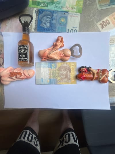 A high-angle, full shot shows several novelty bottle openers shaped like phallic figures and a bottle of liquor laid out on a white surface, with various currencies visible in the background. One bottle opener, shaped as a reclining woman, is attached to the neck of a liquor bottle labeled "Malgrat de Mar - Costa de Barcelona." Another phallic bottle opener with a reclining woman is to the left of the liquor bottle. To the right of the liquor bottle is a single Ukrainian hryvnia banknote. Further to the right, another phallic bottle opener features a reclining woman. Behind the white surface, several banknotes are visible, including Russian rubles and Kyrgyzstani som. The scene is set outdoors, possibly on a table or pavement, with a person's legs and shorts visible in the foreground at the bottom of the image. The lighting suggests it is daytime. There is no indication of the location being in Schwäbisch Hall, Germany.