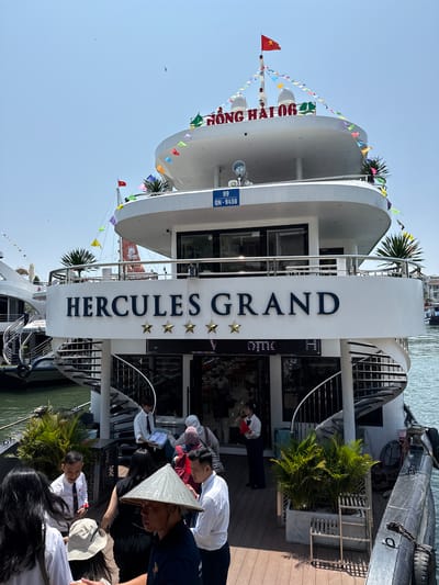 The image depicts the Hercules Grand cruise ship docked in Tuan Chau Ward, Vietnam. The ship's name, "Hercules Grand," is prominently displayed on a vertical sign with three stars underneath. The ship has multiple decks adorned with colorful triangular flags. Passengers, including some wearing traditional conical hats, are boarding the ship.